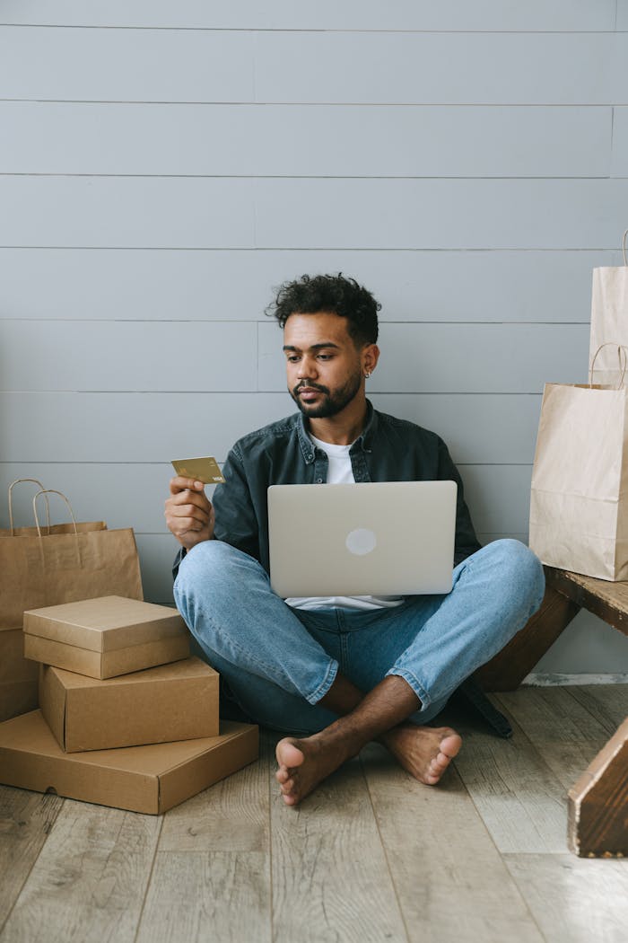Adult male engaged in online shopping with laptop and credit card surrounded by packages.